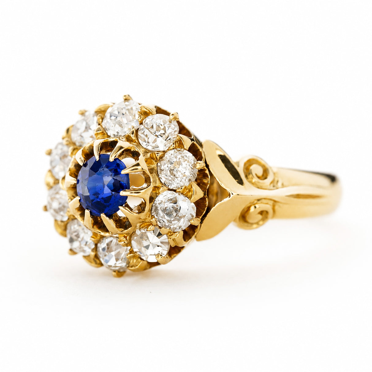 An Antique, Victorian sapphire and old mine-cut diamond cluster engagement ring in yellow gold turned at an angle to show off the shoulders seen against a plain white background.