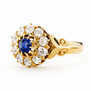 An Antique, Victorian sapphire and old mine-cut diamond cluster engagement ring in yellow gold turned at an angle to show off the shoulders seen against a plain white background.