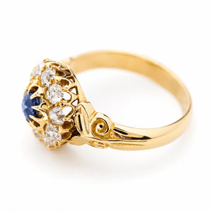 A picture of a Antique, Victorian sapphire and old mine-cut diamond cluster engagement  ring against a white background sat at an angle showing the intricately pierced gallery and scrolling yellow gold shoulders.