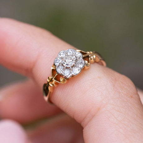 An Antique, Edwardian diamond daisy cluster engagement ring in yellow gold with decorative open-work shoulders shown close-up and worn on a finger.
