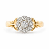 An Antique Edwardian era diamond daisy cluster engagement ring set in yellow gold with ornate scrollwork photographed on a clean white background.