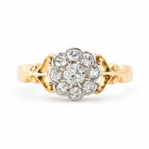 An Antique Edwardian era diamond daisy cluster engagement ring set in yellow gold with ornate scrollwork photographed on a clean white background.