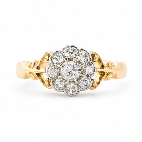 An Antique Edwardian era diamond daisy cluster engagement ring set in yellow gold with ornate scrollwork photographed on a clean white background.