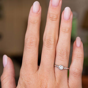 A close-up of a hand wearing a platinum diamond engagement ring with a round-cut central diamond and smaller flanking diamonds.  The ring is Antique from the 1930s Art Deco era. 