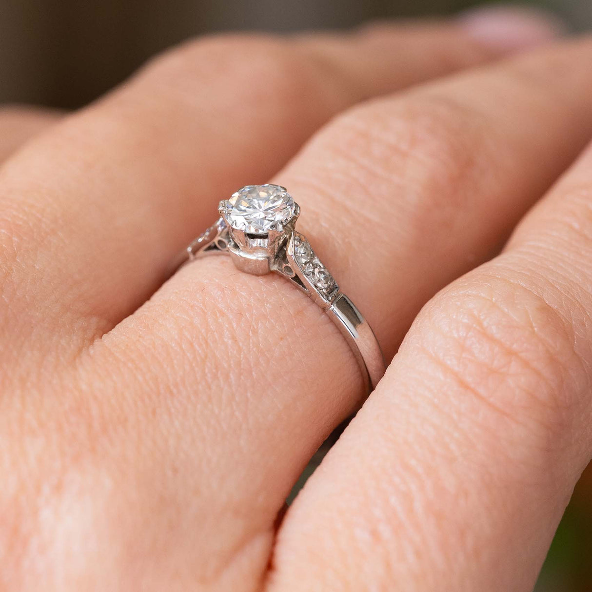 A close-up of a 1930s antique Art Deco diamond engagement ring with a round-cut central diamond and flanking smaller diamonds on either side.   Worn on a person's finger showcasing the platinum setting and band. 
