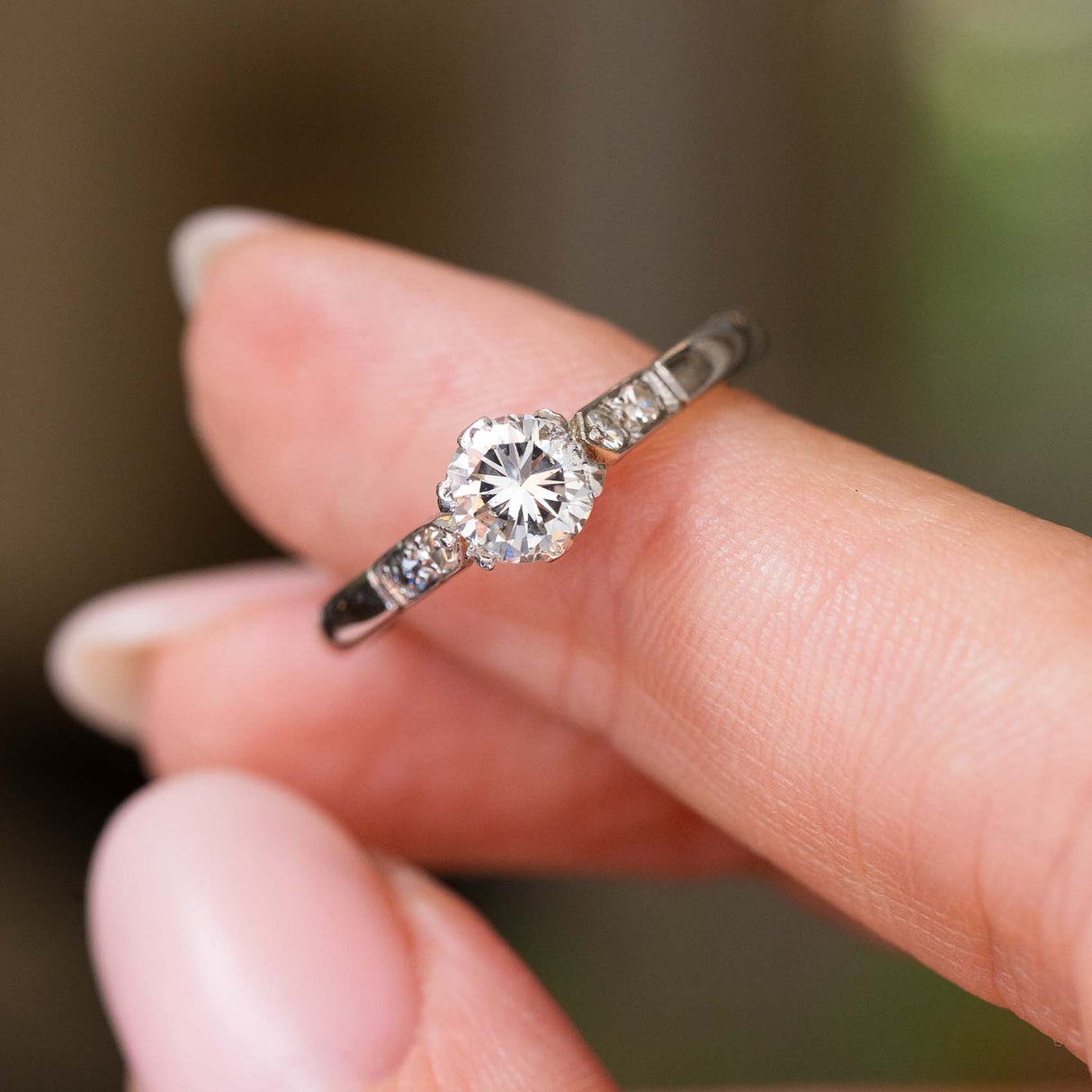 Close up of an Antique, Art-Deco diamond flanked solitaire engagement ring in platinum displayed on a finger against a blurred interior background