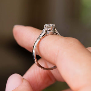 A close-up of an Antique, Art-Deco diamond flanked solitaire engagement ring  in platinum worn on a person's finger, with a soft blurred background.