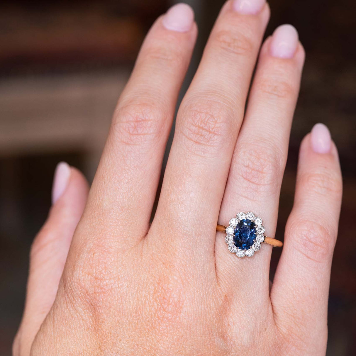 A picture of an Antique, Edwardian blue spinel and diamond coronet cluster engagement ring on a yellow gold band worn on the hand and shown against a blurry background to showcase the proportions of the ring.
