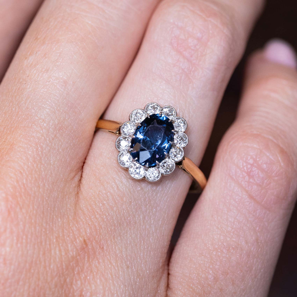 A close-up picture of an Antique, Edwardian blue spinel and diamond coronet cluster engagement ring worn on a finger set on a warm yellow gold band.