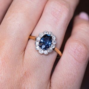 A close-up picture of an Antique, Edwardian blue spinel and diamond coronet cluster engagement ring worn on a finger set on a warm yellow gold band.