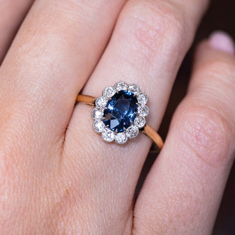 A close-up picture of an Antique, Edwardian blue spinel and diamond coronet cluster engagement ring worn on a finger set on a warm yellow gold band.