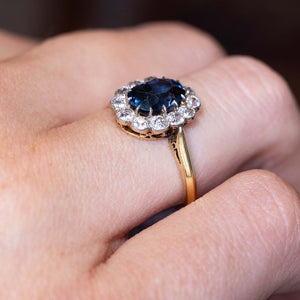 A close-up image of a yellow gold Antique, Edwardian blue spinel and diamond cluster engagement ring in yellow gold worn on a finger and seen from the side to show the shoulders and setting style of the ring.