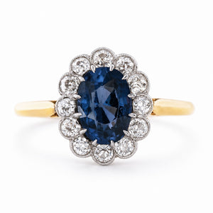an Antique, Edwardian blue spinel oval-cut and diamond cluster engagement ring mounted on a yellow gold band and shown against a white background.