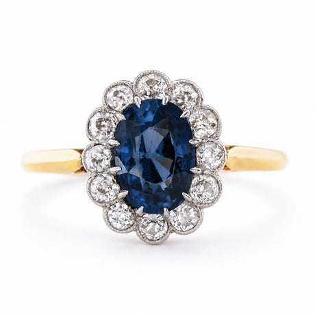 an Antique, Edwardian blue spinel oval-cut and diamond cluster engagement ring mounted on a yellow gold band and shown against a white background.