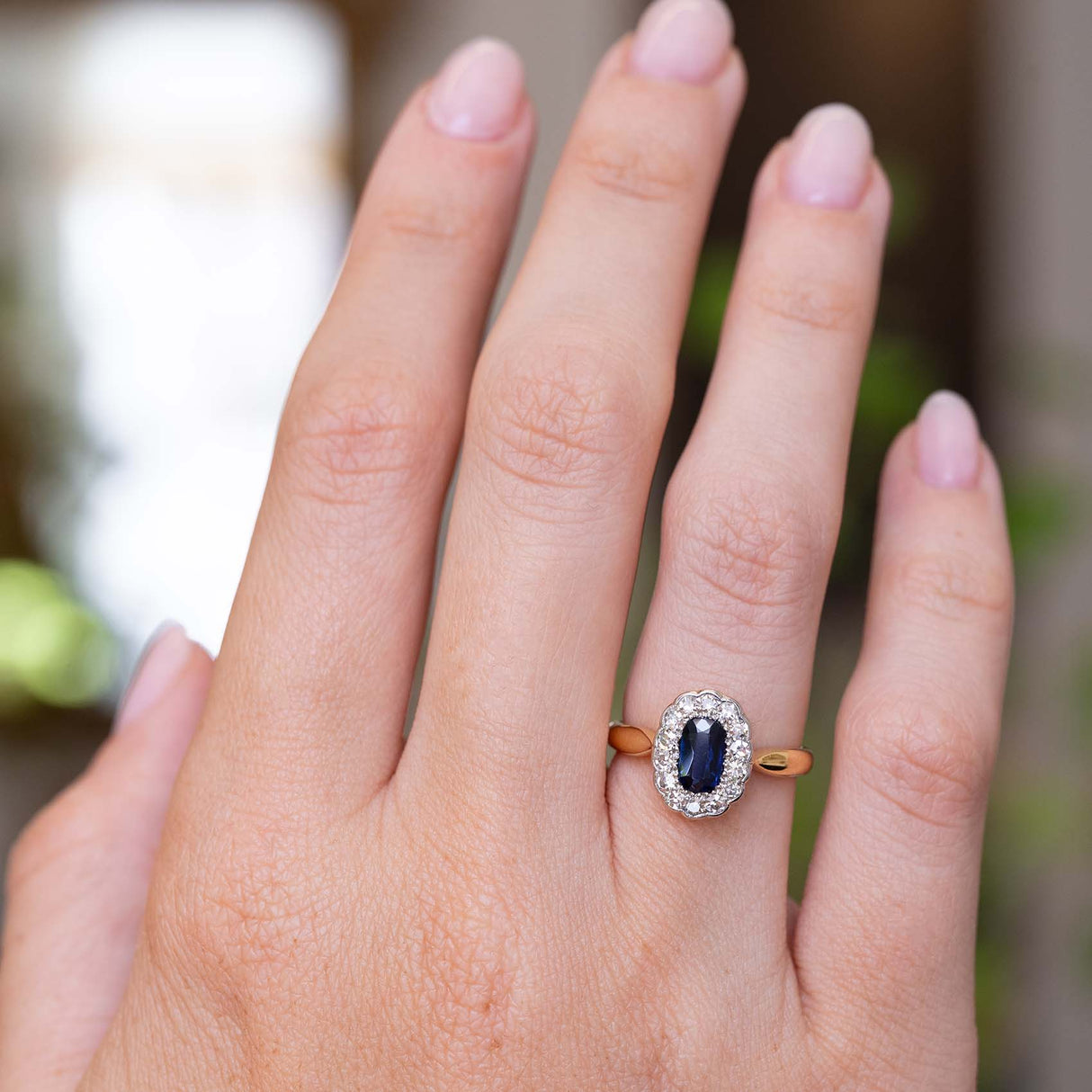 A picture of a sapphire and diamond cluster antique engagement ring on a yellow gold band worn on the finger and shown on the hand to display the proportions of the ring from a distance.