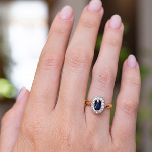 A picture of a sapphire and diamond cluster antique engagement ring on a yellow gold band worn on the finger and shown on the hand to display the proportions of the ring from a distance.