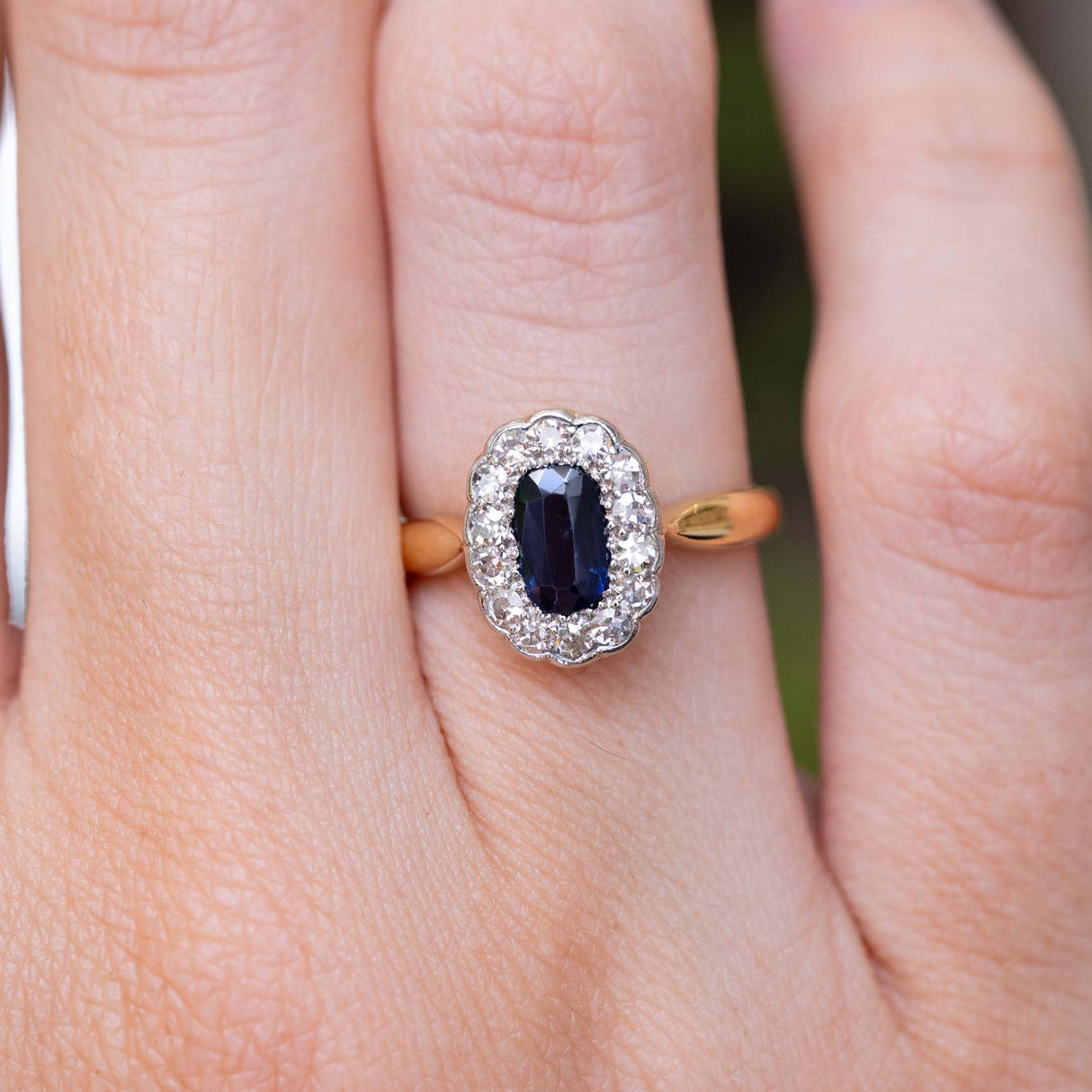 A close-up image of an antique Edwardian cluster ring featuring a sapphire and diamonds on a yellow gold band being worn on the finger.