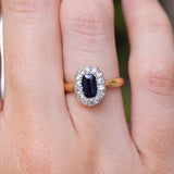 A close-up image of an antique Edwardian cluster ring featuring a sapphire and diamonds on a yellow gold band being worn on the finger.