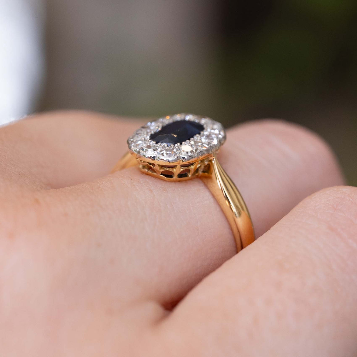 A close-up shot of an Antique Edwardian c.1905 sapphire and diamond cluster engagement ring yellow gold worn on a person's finger and showcased from a low angle to display the rings profile when worn. 