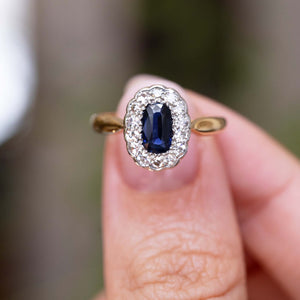 A close-up of a yellow gold antique c.1905 sapphire and diamond cluster engagement ring being held between the thumb and finger and shown against a blurry background. 