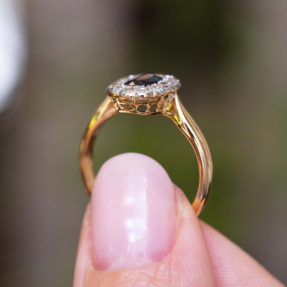 A close-up photograph of an Antique Edwardian sapphire and diamond cluster engagement ring in yellow gold held up and shown from the side to display the pierced open-work gallery of the ring against a blurred background.