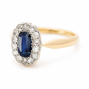 An Antique Edwardian sapphire and diamond cluster engagement ring on a yellow gold band shown from an angle against a white background.