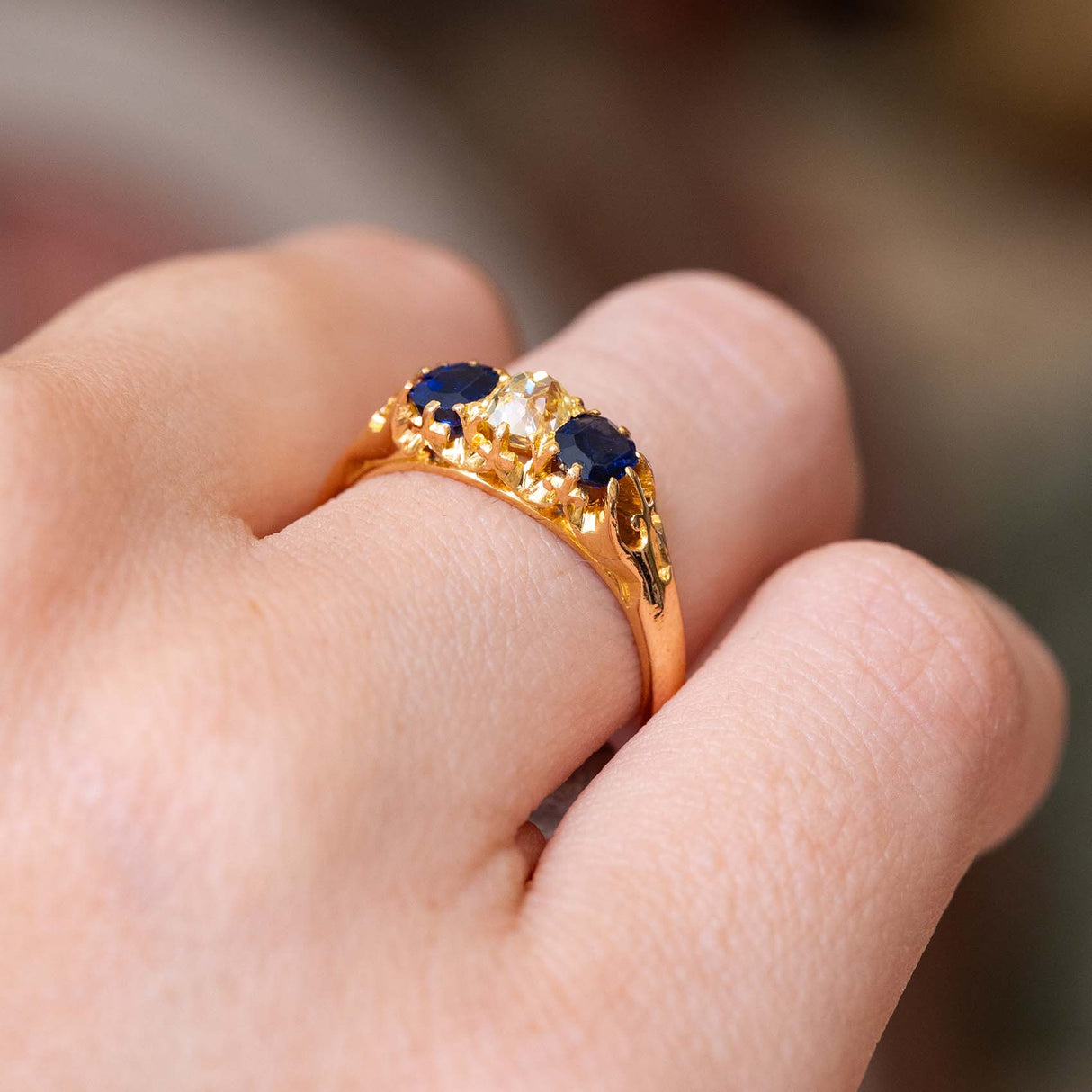 A yellow gold Antique, Victorian sapphire and yellow diamond engagement trilogy ring worn on a person's finger  and shown from the side to display the setting against a softly blurred background.
