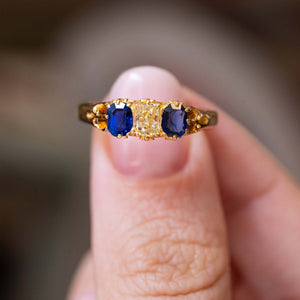 A close-up of a yellow gold Antique, Victorian blue sapphire and cushion-cut yellow diamond three-stone engagement ring held between fingers with a blurred grey background.