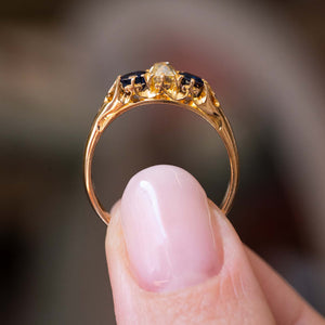 A close-up image of an Antique, Victorian blue sapphire and yellow diamond three-stone engagement ring with an intricately carved gallery held between the fingers and held up to show the ring's profile against a blurred background. 