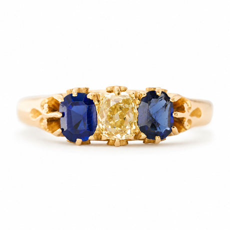 A yellow gold Antique, Victorian blue sapphire and yellow diamond three-stone engagement ring seen against a white background.