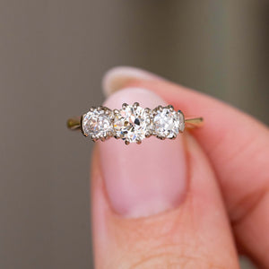 A detailed photo of an yellow gold Antique, Edwardian diamond three-stone engagement ring held in between fingers and shown against a blurry background