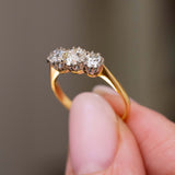 A close-up of a yellow gold Antique, Edwardian diamond three-stone engagement ring held between fingers to show the gallery of the ring against a soft, blurred grey background.