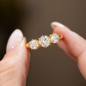 Vintage three-stone engagement ring held between the fingers, with a transitional-cut centre diamond flanked by two round old-cut diamonds.