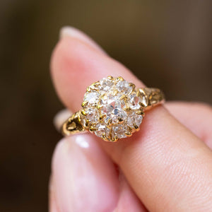 A close-up of an antique Victorian era yellow gold coronet cluster ring balanced on a person's finger against a softly blurred brown background.