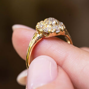 A close-up image of an antique Victorian era yellow gold old-cut diamond coronet cluster engagement ring balanced on the finger and shown from the side to showcase the ring's gallery and settings seen against a blurry background.