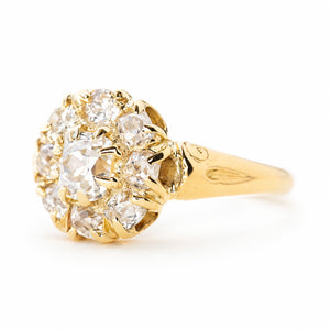 An antique yellow gold cluster engagement ring with old cut diamonds and engraved shoulders shown from the side against a plain white background. 