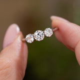 A close-up of an Antique, Edwardian diamond three-stone engagement ring in platinum held between fingers against a soft, blurred green and brown background.