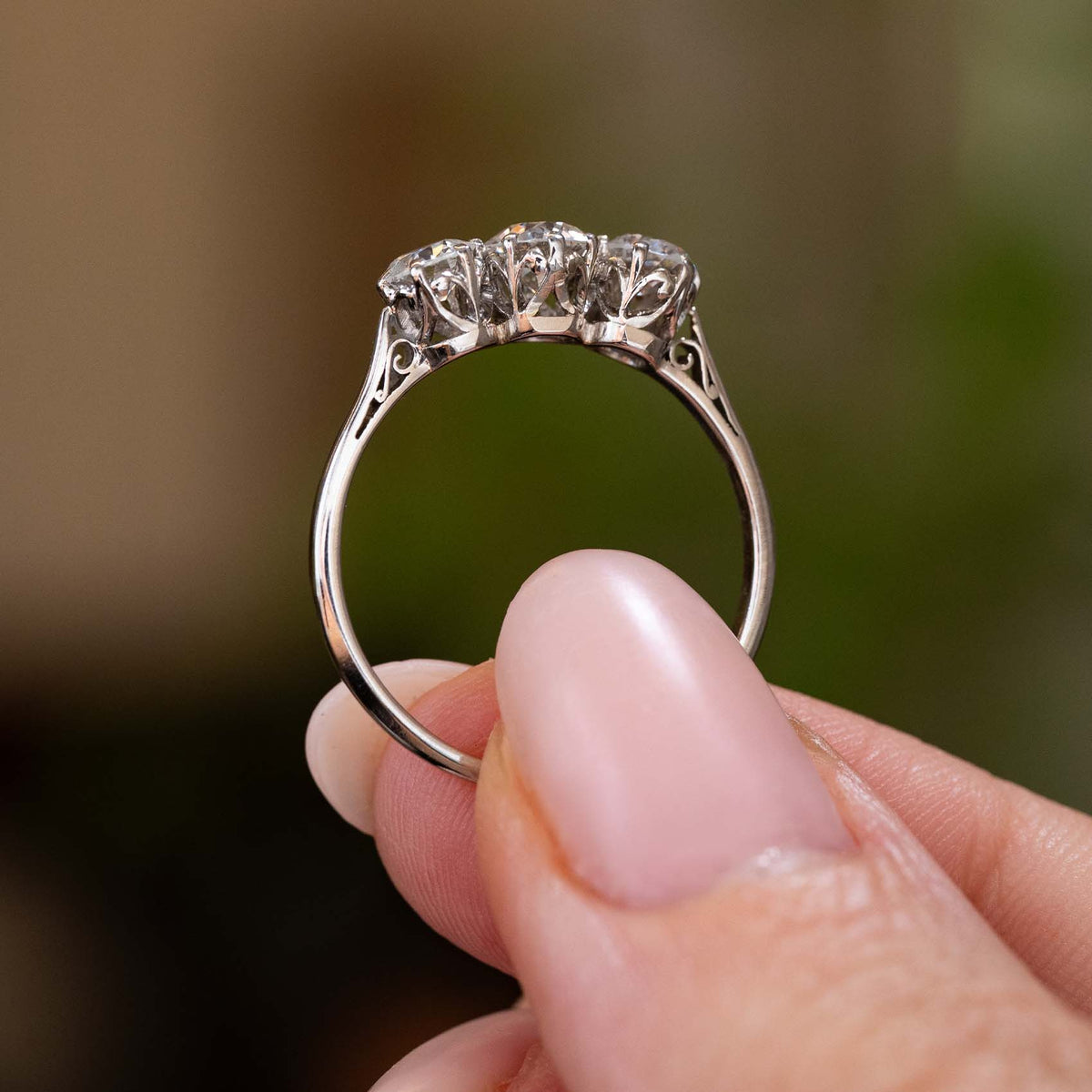 A close-up of a platinum Antique, Edwardian diamond three-stone engagement rings held between fingers against a soft, blurred green and brown background. The ring is held to display its profile and intricate setting style.