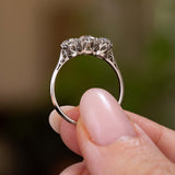 A close-up of a platinum Antique, Edwardian diamond three-stone engagement rings held between fingers against a soft, blurred green and brown background. The ring is held to display its profile and intricate setting style.