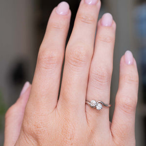 A photo of a hand wearing an Antique, Edwardian toi et moi style two stone diamond engagement ring in platinum against a softly blurred background, showing the proportions of the ring when worn.