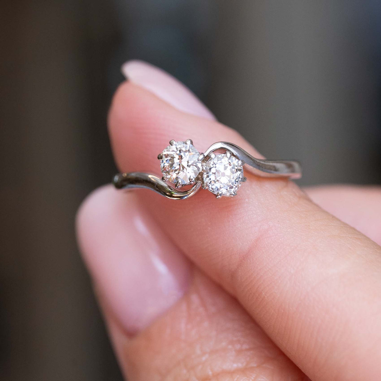 A close-up of an Antique, Edwardian two stone toi et moi diamond engagement ring in platinum held between the fingers against a blurry background. 