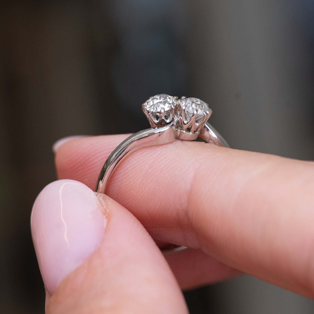 A close-up picture of a platinum Antique, Edwardian toi et moi diamond engagement ring held between fingers at an angle showcasing the ring's profile against a blurred dark background.