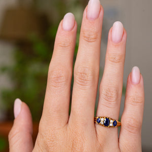 A close-up of a hand wearing an Antique, Edwardian blue sapphire and diamond five-stone engagement ring in yellow gold set against a softly blurred background.