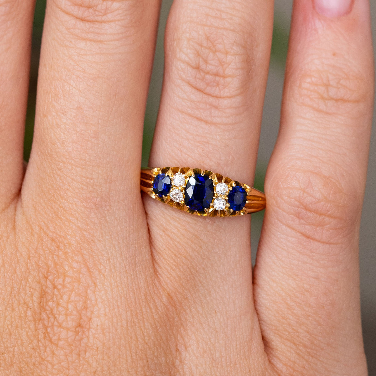 A close-up of an Antique, Edwardian sapphire and diamond five-stone engagement ring.  Featuring three cushion-cut blue sapphires interspersed with old-cut diamonds on a yellow gold band, worn on a finger.