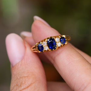 A yellow gold antique Edwardian five-stone engagement ring set with three cushion-cut blue sapphires and smaller accent diamonds, held between fingers against a blurred green background.