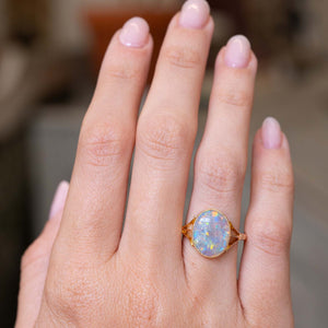 A close-up picture of an outstretched hand wearing a gold ring with an oval opal gemstone in yellow gold mount displayed at a distance to showcase the scale of the ring when worn.