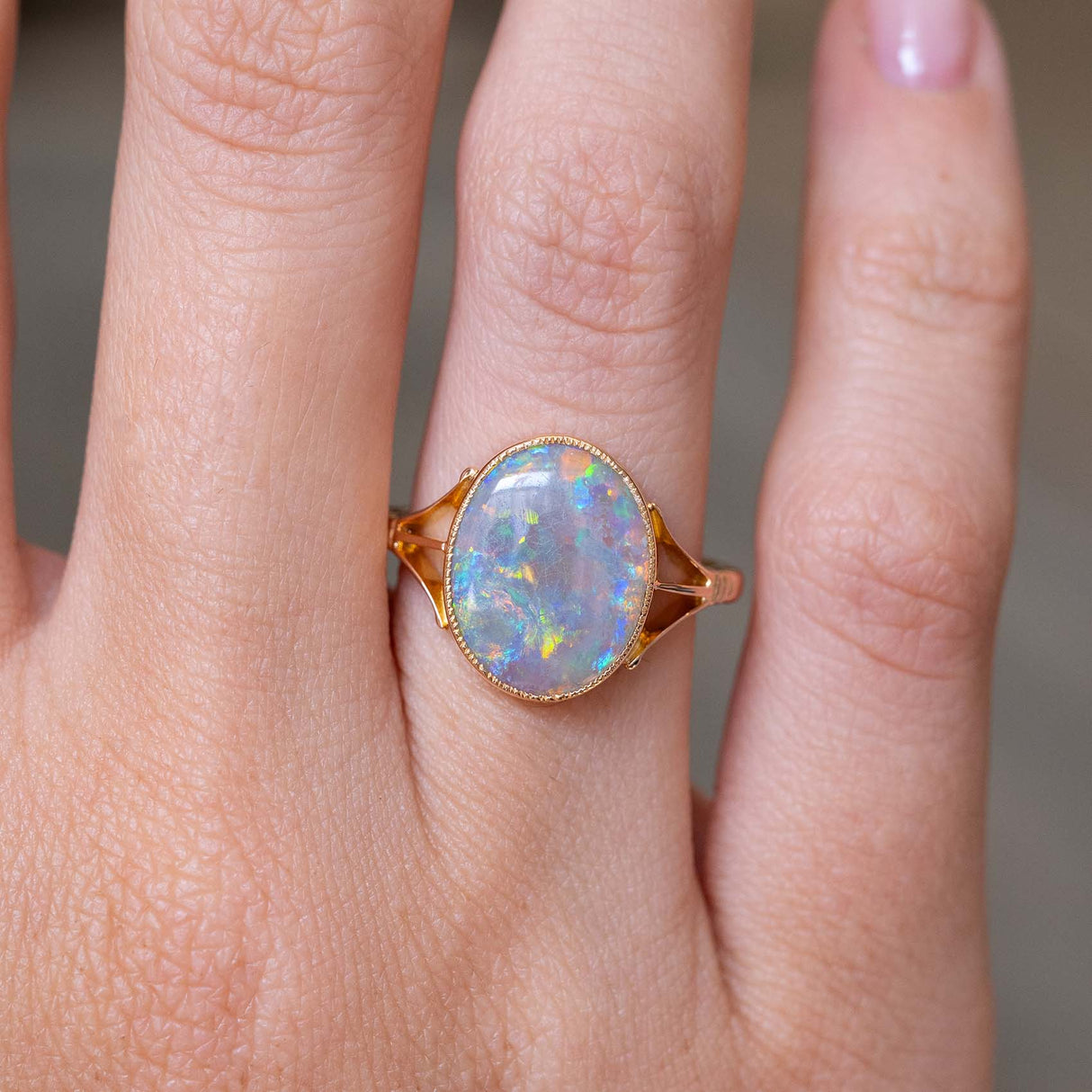 A close up image of an antique opal solitaire ring with decorative tri-split shoulders and an intricate bezel setting style against a blurry background.