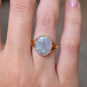A close up image of an antique opal solitaire ring with decorative tri-split shoulders and an intricate bezel setting style against a blurry background.