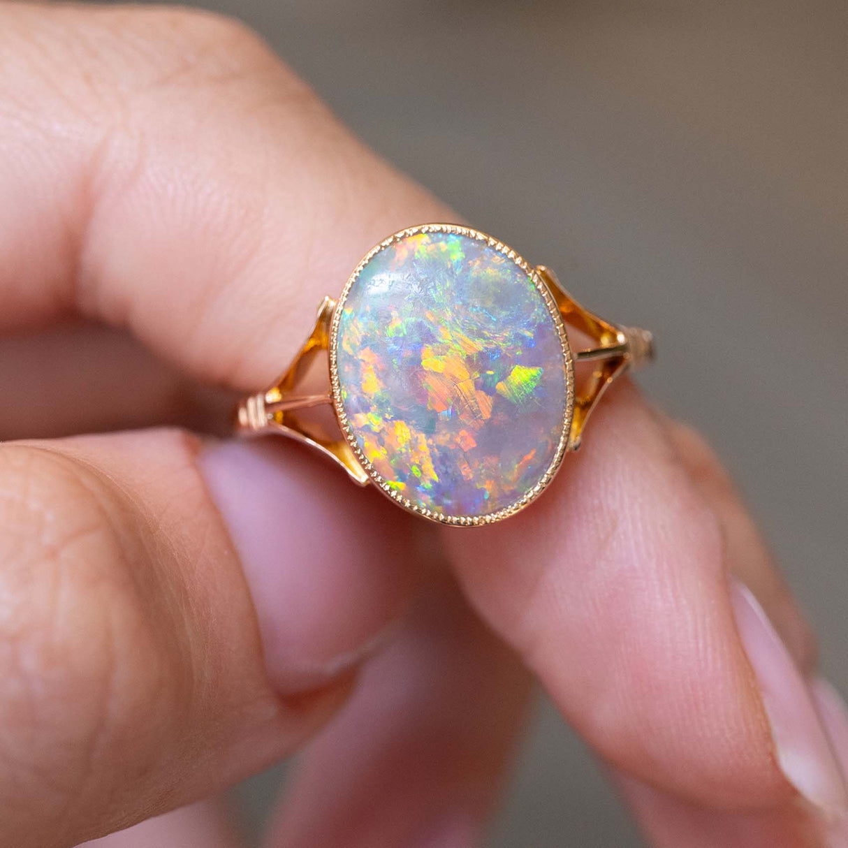 A close-up image of an antique opal solitaire ring in yellow gold and made in the Edwardian era, placed between the finger and thumb against a blurred grey background. 