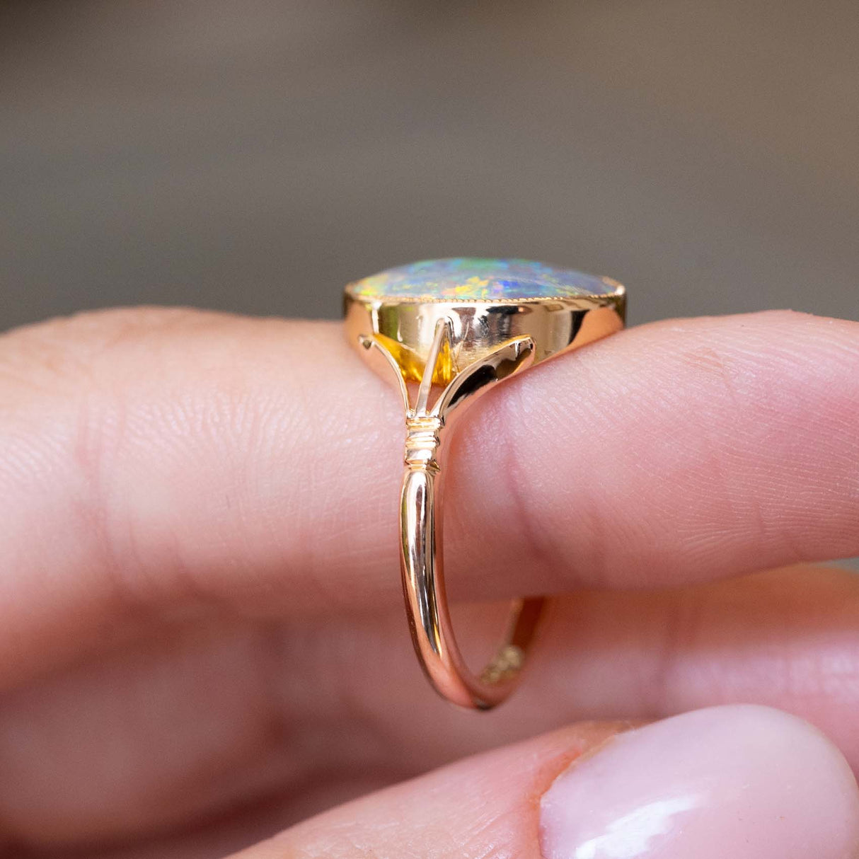 A close-up of an antique, Edwardian era opal solitaire ring with decorative split shoulders mounted in yellow gold seen on a finger to display the band and its details.
